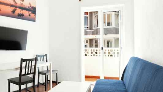 Interiørbilde av stue med en blå sofa, et hvitt bord, to svarte stoler og en TV på veggen. Bak glassdøren kan man se en bygning. (AI-generert tekst)