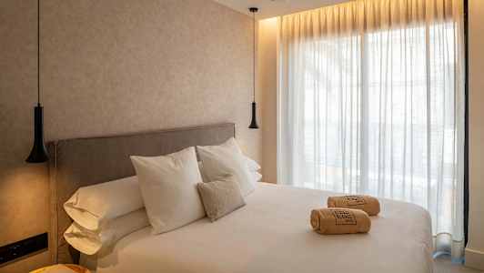 Et hotellrom med en stor seng, hvitt sengetøy og puter. På sengen ligger sammenrullede håndklær. Vegger og gardiner er lyse, og rommet har hengende lamper. (AI-generert tekst)