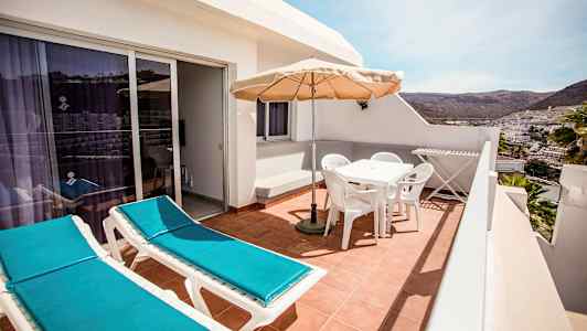 En terrasse med to solsenger, utemøbler med parasoll og utsikt over en by og fjell i det fjerne. (AI-generert tekst)
