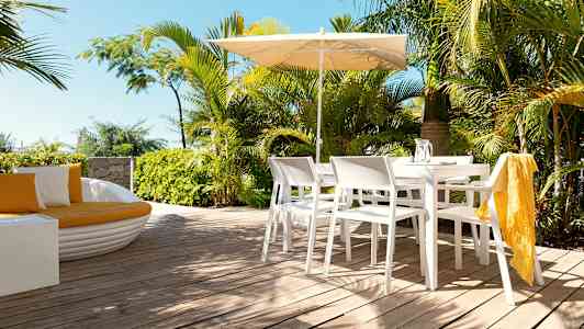 En uteplass med hvite stoler og bord under en parasoll, omgitt av grønne planter. (AI-generert tekst)