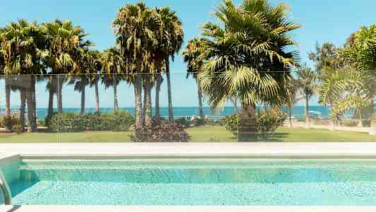 Utsikt fra et basseng mot en hage med palmer og havet i bakgrunnen. (AI-generert tekst)