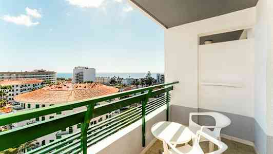 Balkong med bord og stoler samt utsikt over bygninger og havet. (AI-generert tekst)