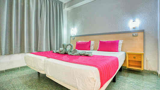 Seng med rosa teppe og puter i et rom med grå gardiner og en trebenk. (AI-generert tekst)