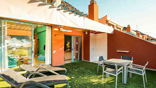 En terrasse med solsenger, bord og stoler samt utsikt inn mot et soverom. (AI-generert tekst)