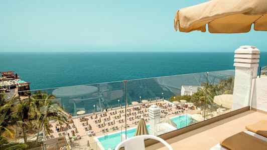 Utsikt fra en terrasse med bord og stoler utover et basseng omgitt av solsenger og parasoller, med utsikt over sjøen i bakgrunnen. (AI-generert tekst)