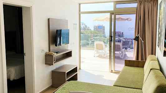 En hotellromutsikt med en grønn sofa og en TV på veggen, med en døråpning til et soverom og en terrasse som viser omgivelsene utenfor. (AI-generert tekst)