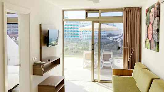 Et lyst hotellrom med sofa, veggmontert TV og skyvedører i glass som fører ut til en møblert balkong. (AI-generert tekst)