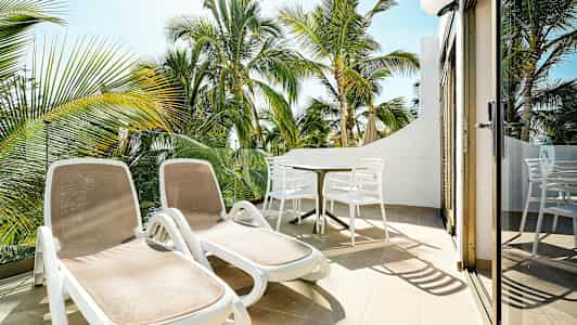 Solstoler og spisebord på en terrasse med utsikt mot palmetrær. (AI-generert tekst)