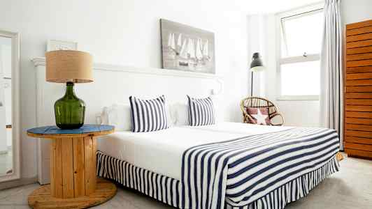 Et soverom med en seng kledd i stripete tekstiler, et bord med en grønn glasslampe og en rottingstol ved siden. (AI-generert tekst)