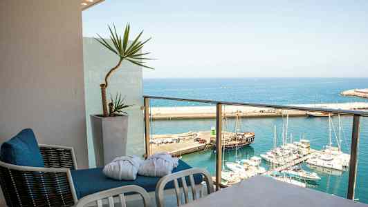 Utsikt over en havn med flere båter fra en balkong med stoler og en potteplante. (AI-generert tekst)