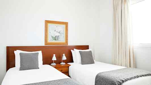 To enkeltsenger med grå pledd og puter, plassert ved siden av en trefarget sengegavl. Et innrammet portrett henger på veggen over sengegavlen. (AI-generert tekst)