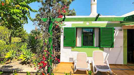 Bilde av et hus med grønne skodder og en terrasse med to solsenger. Beplantning med blomster og planter omgir området. (AI-generert tekst)