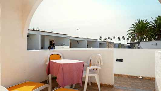 Den viser en uteplass med solsenger og et bord med stoler. Bakgrunnen har små hus og palmer. (AI-generert tekst)