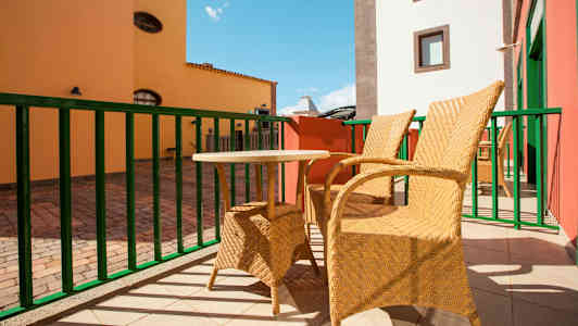 To flettede stoler og et lite bord står på en solfylt terrasse med fargerike bygninger i bakgrunnen. (AI-generert tekst)