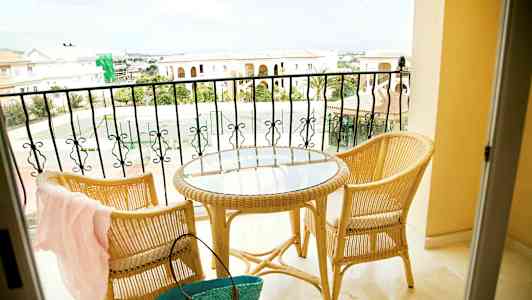 To rottingstoler og et rundt bord med glassplate står på en balkong med utsikt over steinbygninger og et tennisområde. (AI-generert tekst)