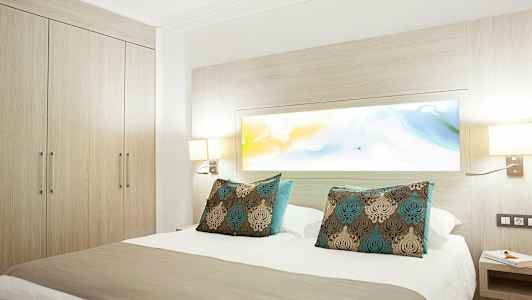 Hotellrom med en dobbeltseng og dekorative puter, omringet av lyst treverk og lamper. (AI-generert tekst)