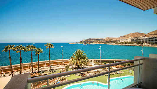 Utsikt fra en balkong over et bassengområde og en kystlinje med palmer, havet og bygninger i det fjerne. (AI-generert tekst)