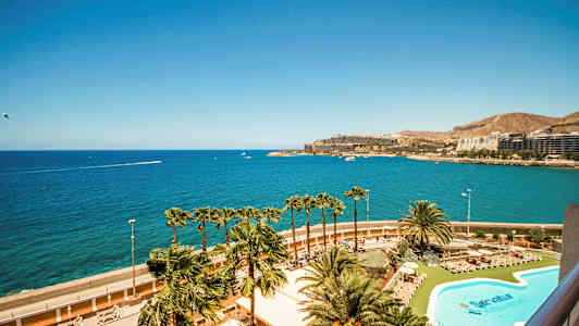 Utsikt over et basseng og palmer med havet og bygninger i bakgrunnen. (AI-generert tekst)