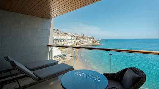 Utsikt fra en balkong mot havet og en strand, med bord og stoler samt solsenger i forgrunnen. (AI-generert tekst)