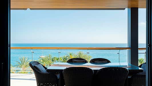 Bilde av en terrasse med bord og stoler, havutsikt og palmer i bakgrunnen. (AI-generert tekst)