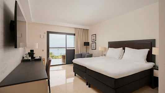 Et hotellrom med en stor seng, et skrivebord og en TV. Gjennom glassdøren sees en balkong og utsikt mot havet. (AI-generert tekst)