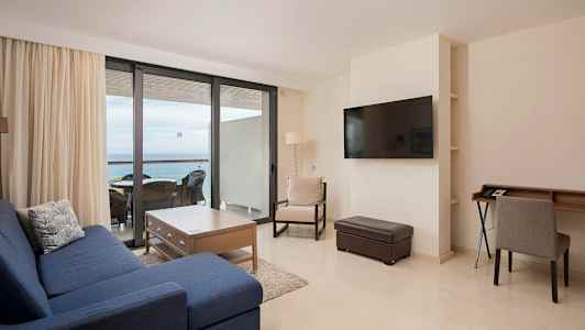 En stue med blå sofa, trebord og stoler ved et stort vindu mot balkongen. (AI-generert tekst)