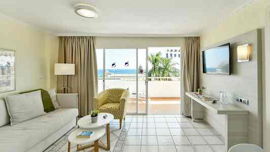 En stue med lyse møbler og en stor glassdør som fører til en balkong med utsikt over havet. (AI-generert tekst)