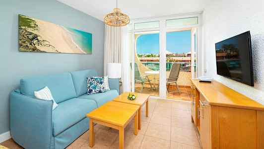 Et oppholdsrom med en blå sofa, to trebord, en TV på en skjenk og utsikt over en terrasse med utemøbler. (AI-generert tekst)