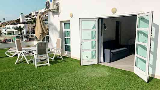 Utendørsområde med grønt gress, sittegruppe og åpne dører til et innendørsområde. (AI-generert tekst)