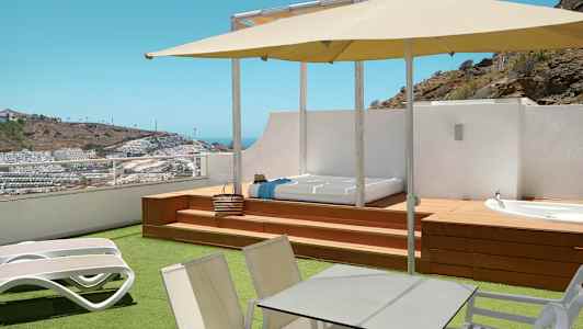 Terrasse med solsenger, bord, parasoll og utsikt over et kupert område med bygninger. (AI-generert tekst)