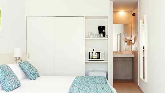 Et hotellrom med en seng, lamper og et minikjøkken som inkluderer en mikrobølgeovn og en kaffemaskin. (AI-generert tekst)