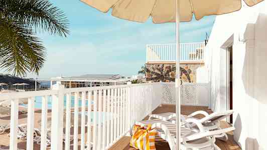 Utsikt over en terrasse med solsenger, parasoll og en strandveske. I bakgrunnen vises et basseng og palmer. (AI-generert tekst)