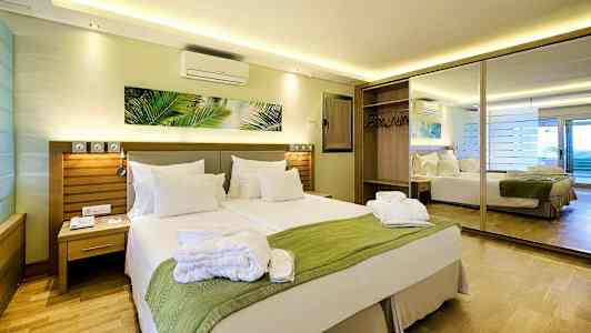 Et hotellrom med en dobbeltseng, et grønt teppe og hvite puter, samt et speil og garderobe i bakgrunnen. (AI-generert tekst)