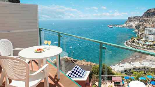 Utsikt fra balkong med bord og to stoler, hvor glass er servert, mot havet og kystlandskap. (AI-generert tekst)