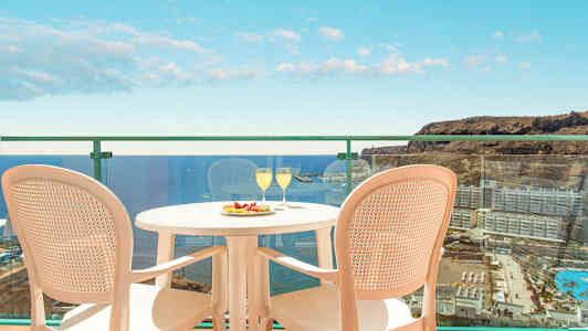 Bilde av en balkong med utsikt over havet og omkringliggende bygninger. På balkongen står et bord med to glass og en tallerken med frukt samt to stoler. (AI-generert tekst)