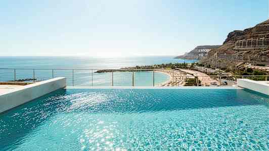 Utsikt over et infinitybasseng med solskinn på vannoverflaten og en sandstrand i bakgrunnen. (AI-generert tekst)