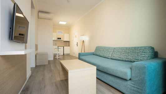 En stue med en blå sofa og et bord foran, samt en TV på veggen og et integrert kjøkken i bakgrunnen. (AI-generert tekst)