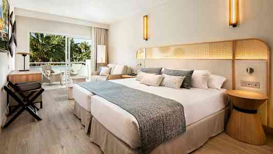 Et hotellrom med en stor dobbeltseng, sittegruppe og balkong med utsikt mot grønne palmer. (AI-generert tekst)