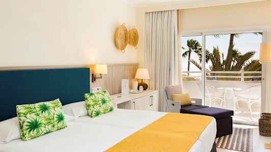 Et hotellrom med en dobbeltseng med gult sengeteppe og grønne pynteputer, lys innredning og en balkong med utsikt mot palmer og havet. (AI-generert tekst)