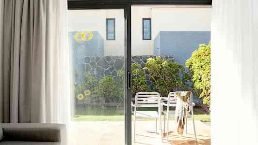 Bilde av en uteplass med to hvite stoler og et bord, omgitt av planter og en steinmur, sett gjennom en glassdør. (AI-generert tekst)