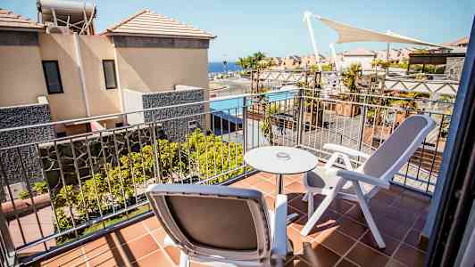 En balkong med to stoler og et bord, utsikt mot bygninger og et basseng. (AI-generert tekst)