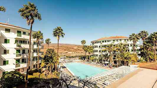 Bilde av et hotellområde med et stort utendørsbasseng omgitt av solsenger. To hvite bygninger med grønne vinduer har palmer i forgrunnen. (AI-generert tekst)