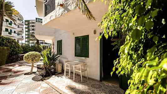 En uteplass med bord og stoler foran en hvit bygning med grønne skodder. Omgitt av trær og busker. (AI-generert tekst)