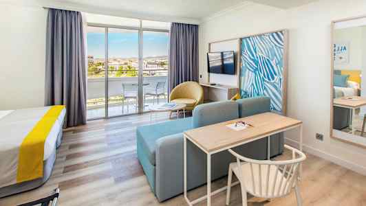 Et hotellrom med seng, skrivebord, sofa, TV og glassdører som fører til en balkong. (AI-generert tekst)