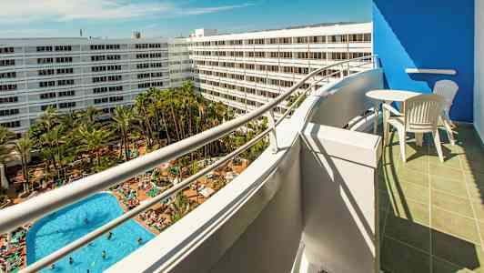 Utsikt fra en balkong mot et bassengområde med mange solsenger og grøntområder, omgitt av hvite hotellbygninger. (AI-generert tekst)