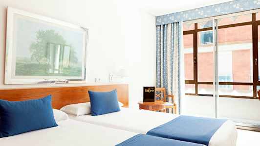 Et hotellrom med to enkeltsenger som har blåe tepper og puter, en stol ved et bord samt et vindu med blått gardin. (AI-generert tekst)