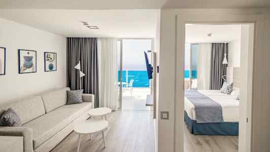 Stue med sofa og bord ved siden av soverommet med dobbeltseng. Havutsikt fra balkongen. (AI-generert tekst)