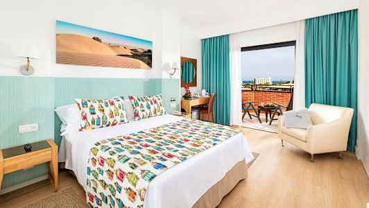 Hotellrom med dekorativt sengeteppe, lenestol og skrivebord samt utsikt fra balkongen. (AI-generert tekst)