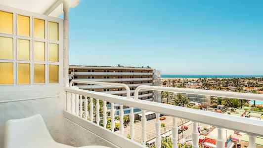 Utsikt fra en balkong med sittemøbler, mot en bygning og havet i bakgrunnen. (AI-generert tekst)