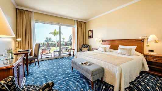 Et hotellrom med en dobbeltseng, skrivebord, stol og store glassdører som fører ut til en frodig terrasse. (AI-generert tekst)